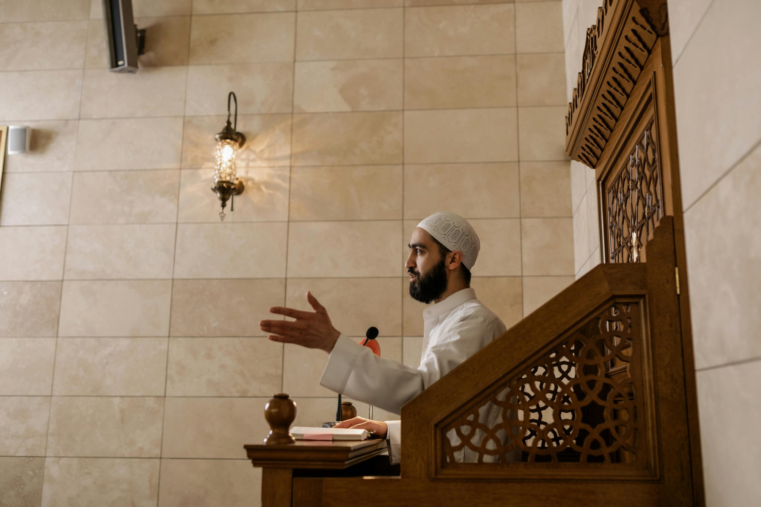 An imam delivering a sermon from a wooden lectern inside a beautifully decorated mosque.